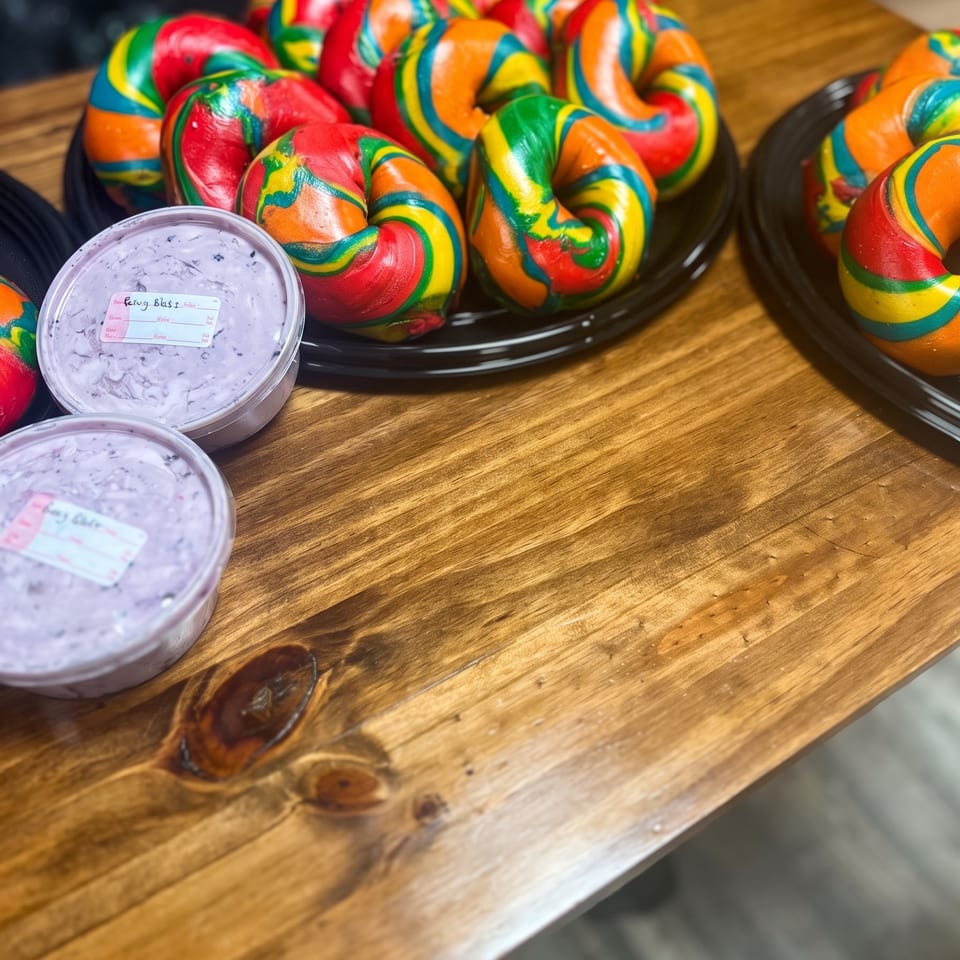 Colorful rainbow bagels arranged on black plates beside tubs of lavender cream cheese on a wooden table.