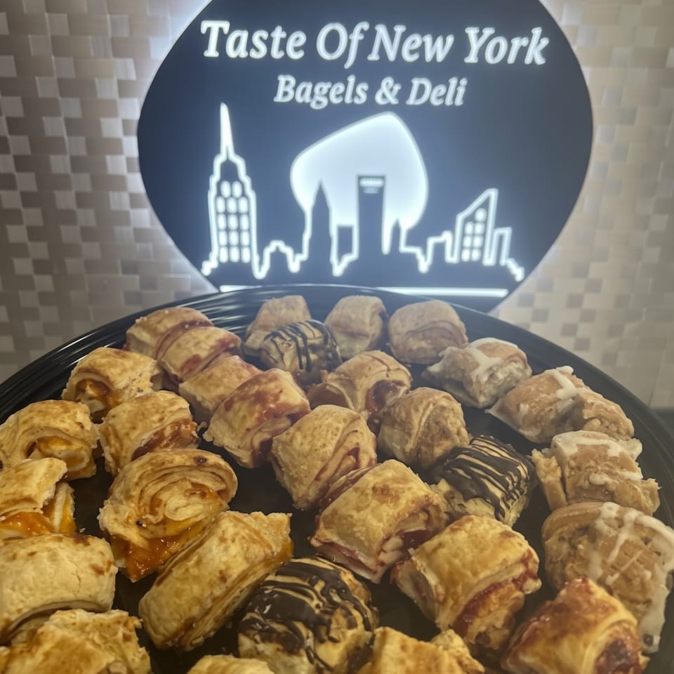 Platter of assorted golden pastries, some drizzled with chocolate, displayed beneath a Taste of New York Bagels & Deli sign.