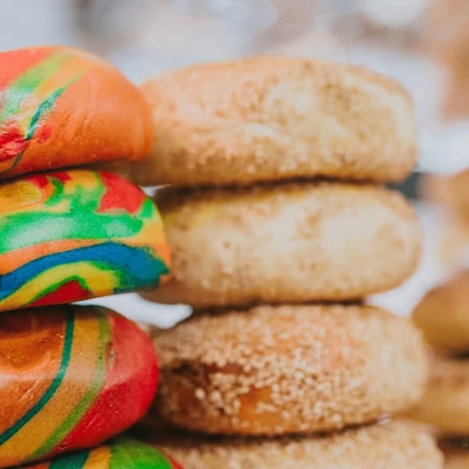 Stacks of colorful rainbow bagels alongside plain sesame bagels in a bakery display.