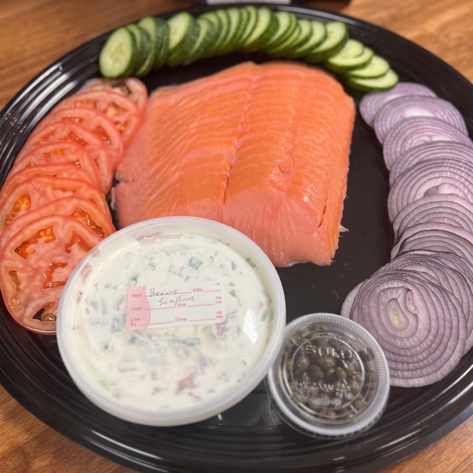 Black platter of sliced salmon, tomatoes, cucumbers, red onions, scallion cream cheese, and olives on a wooden table.
